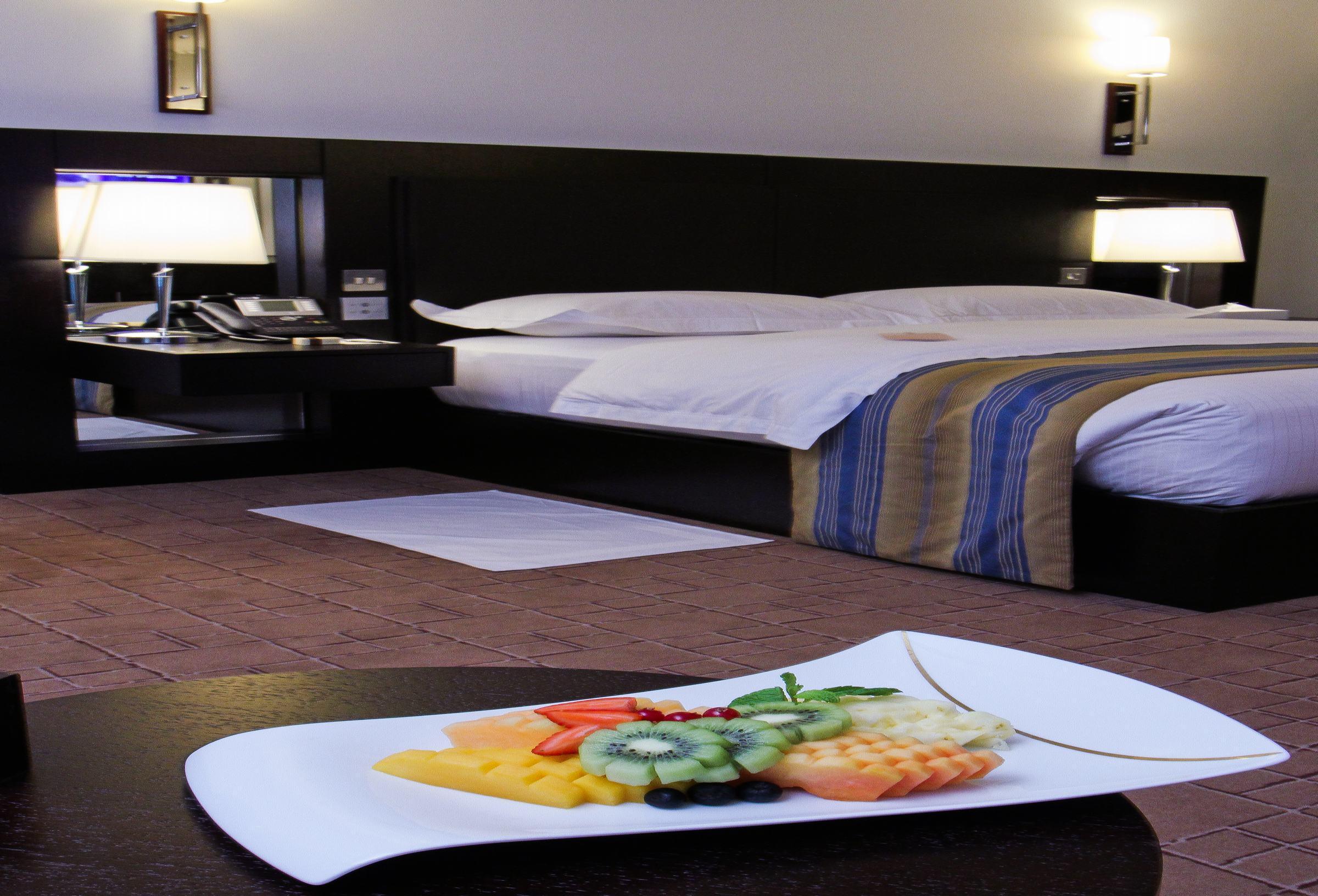Dubai International Terminal Hotel Exterior foto The photo shows a modern hotel room with a large bed covered in white linens and a blue-striped throw. On either side of the bed, there are bedside tables with lamps. In the foreground, there is a decorative platter of assorted fresh fruits, includin