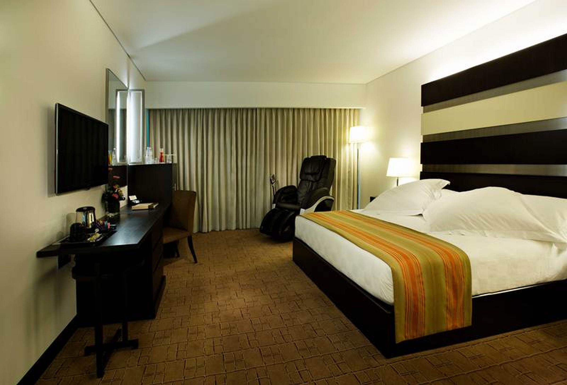 Dubai International Terminal Hotel Exterior foto The photo shows a hotel room with a modern design. There is a large bed with white bedding and an orange-accented throw blanket. A comfortable chair is positioned in the corner next to a floor lamp. The room features a television mounted on the wall 