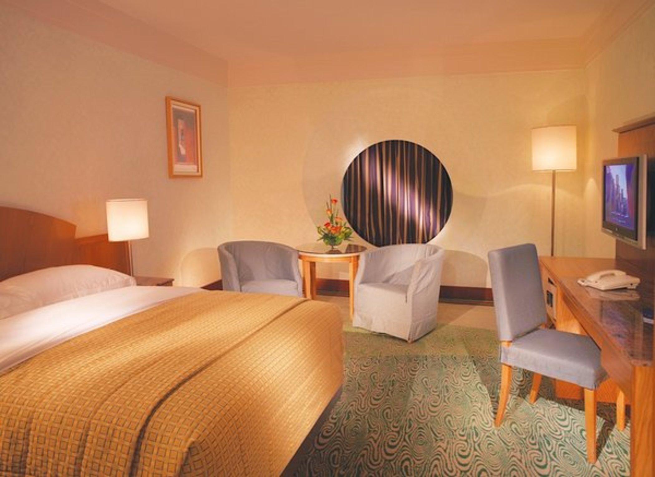Dubai International Terminal Hotel Quarto foto The photo shows a cozy hotel room featuring a large bed with a patterned yellow bedspread. There are two upholstered chairs and a small table beside them. A round window is set into the wall, partially covered by curtains. A television is mounted on 