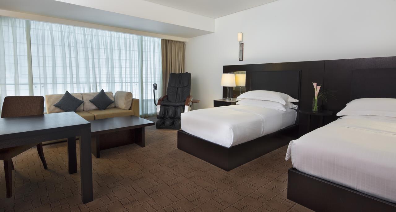 Dubai International Terminal Hotel Exterior foto The photo shows a modern hotel room featuring two twin beds, a small table, and a seating area with a couch. There is a large window with sheer curtains allowing natural light to enter, and a chair positioned nearby. The room has a minimalist design 