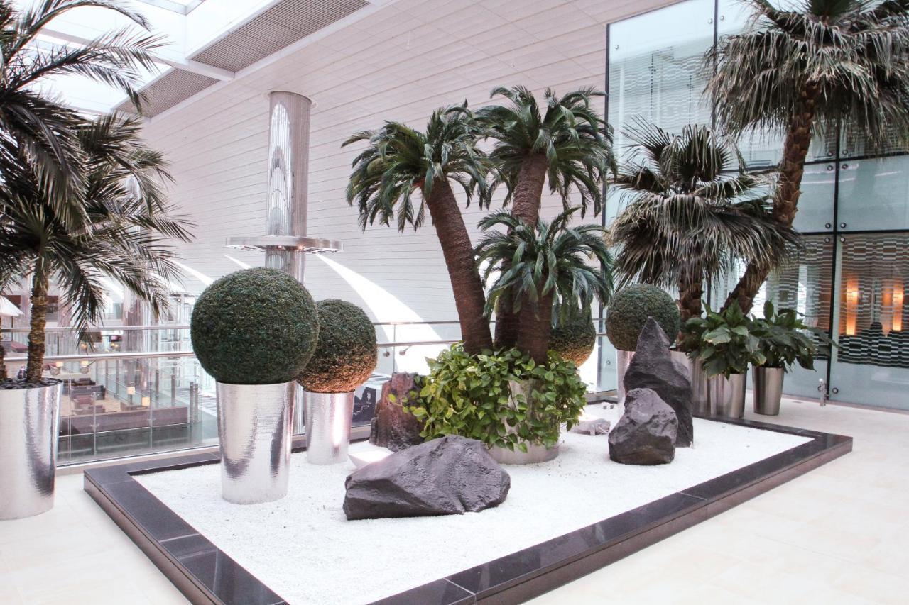 Dubai International Terminal Hotel Exterior foto The photo showcases a modern indoor landscaping setup. It features several tall palm trees surrounded by decorative plants and shrubs. There are large, rounded topiary plants in silver planters, as well as natural rocks scattered around a white grave