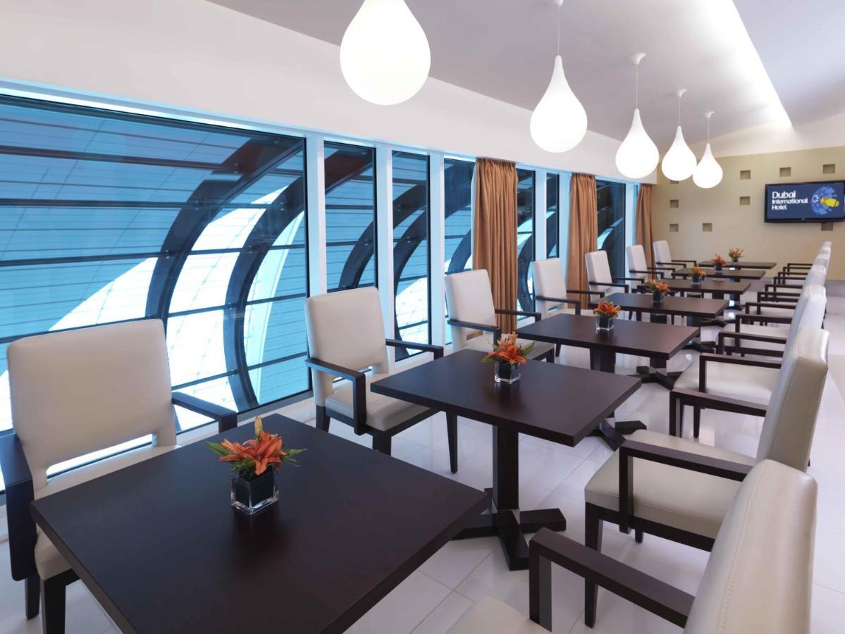 Dubai International Terminal Hotel Exterior foto The photo shows a modern restaurant or dining area with several tables and chairs arranged neatly. Each table is dark wood with a small potted plant as a centerpiece. The seating consists of light-colored upholstered chairs. Large windows provide nat